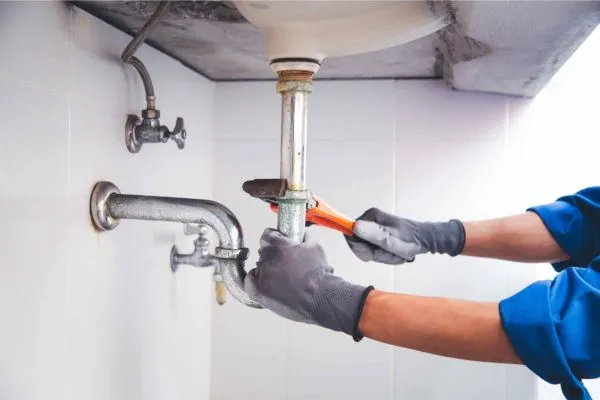 Close-up of a plumber's hands in grey work gloves using an orange pipe wrench to tighten a chrome p-trap under a sink.