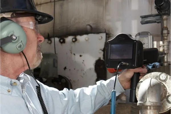 An industrial technician wearing a hard hat and noise-canceling headphones uses a handheld ultrasonic detector to inspect metal pipes for pressurized leaks.